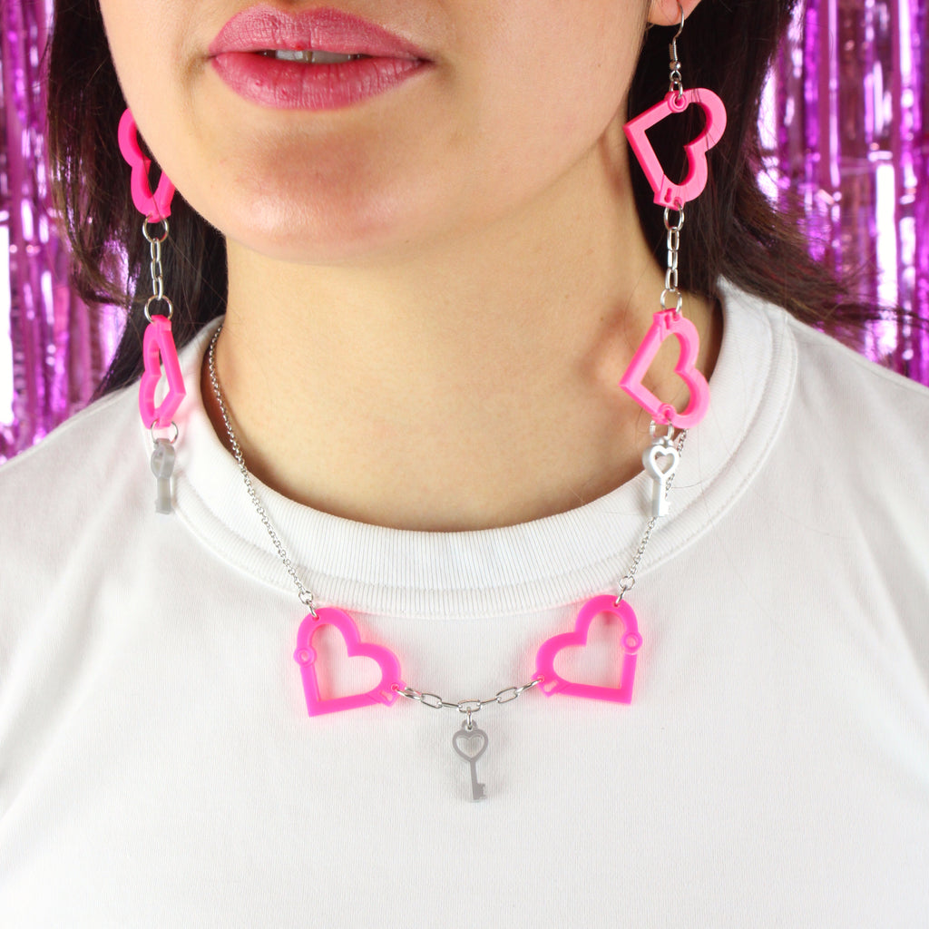 Model wearing neon pink acrylic heart charms chained together to resemble handcuffs, with a key charm in the middle of the two handcuff pendants, on a stainless steel chain. Model is also wearing the matching earrings.