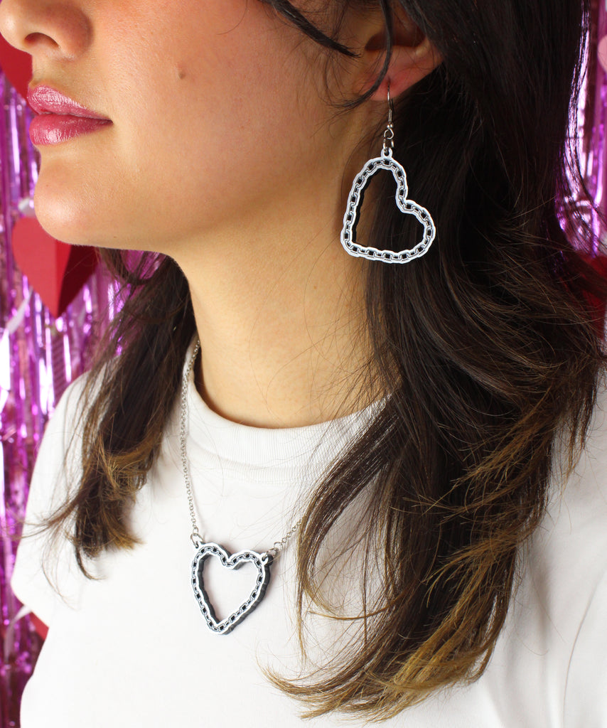 Model wearing a stainless steel chain with a cut out heart pendant with a chain style outline with matching earrings