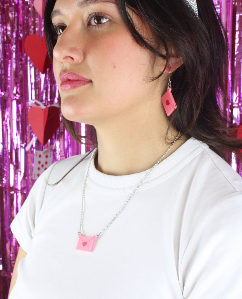 Model wearing pink envelope pendant sealed with a red heart on a stainless steel chain, with the matching earrings.