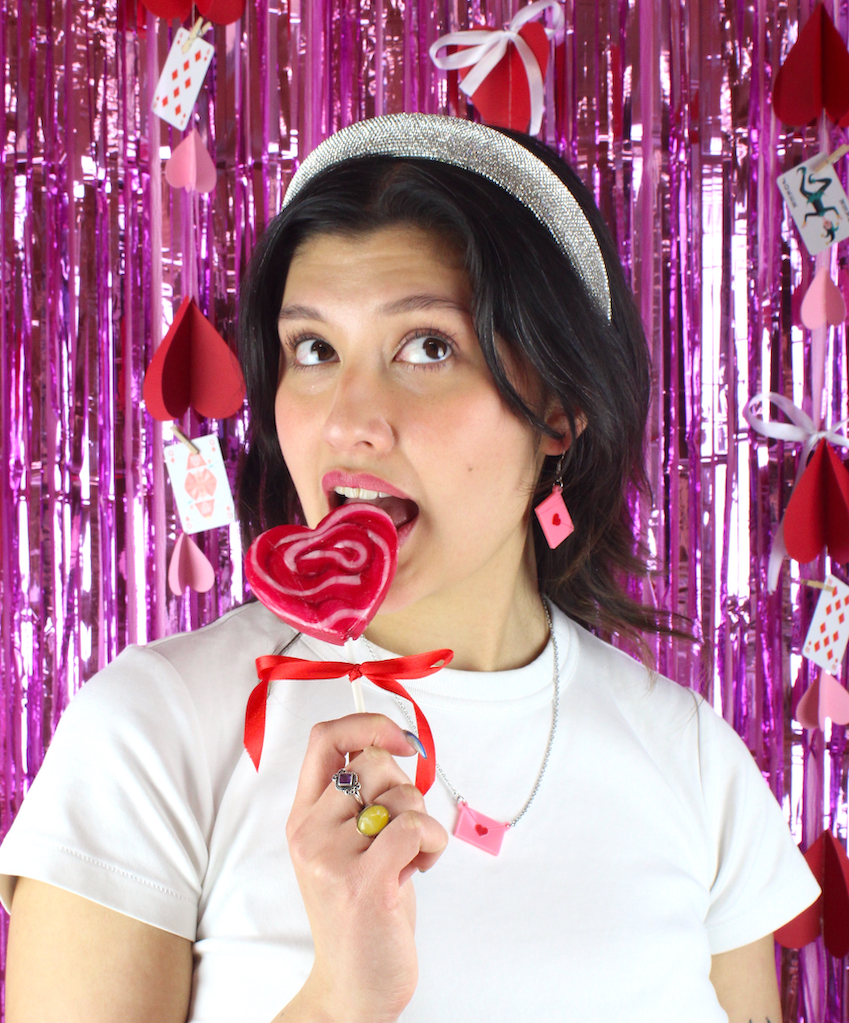 A girl with brown hair licking a heart-shaped lollipop with perspex pink heart envelope earrings and necklace set