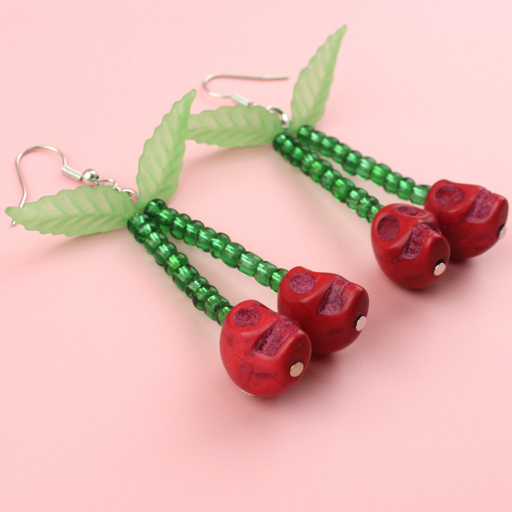 Red skull cherry charms on stems made out of green beads with two leaves attached at the top on stainless steel earwires