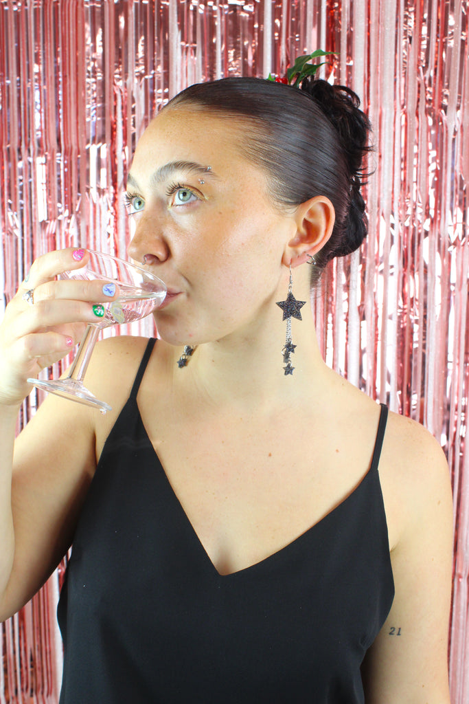 Model wearing a black glitter acrylic star with three chains dangling from it with mini black glitter acrylic stars at the end of each chain on stainless steel earwires, the model is drinking frm a cocktail glass