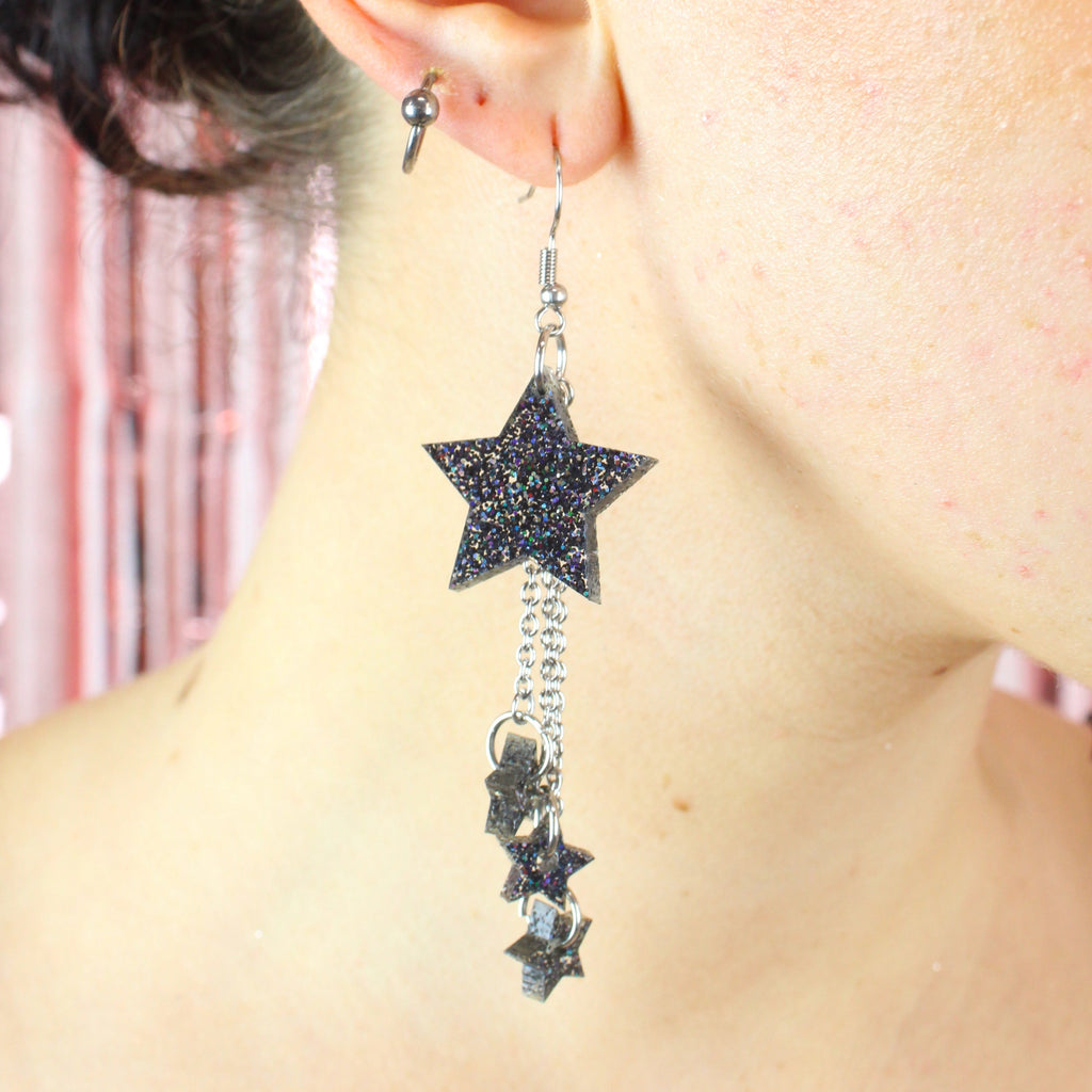 Model wearing a black glitter acrylic star with three chains dangling from it with mini black glitter acrylic stars at the end of each chain on stainless steel earwires