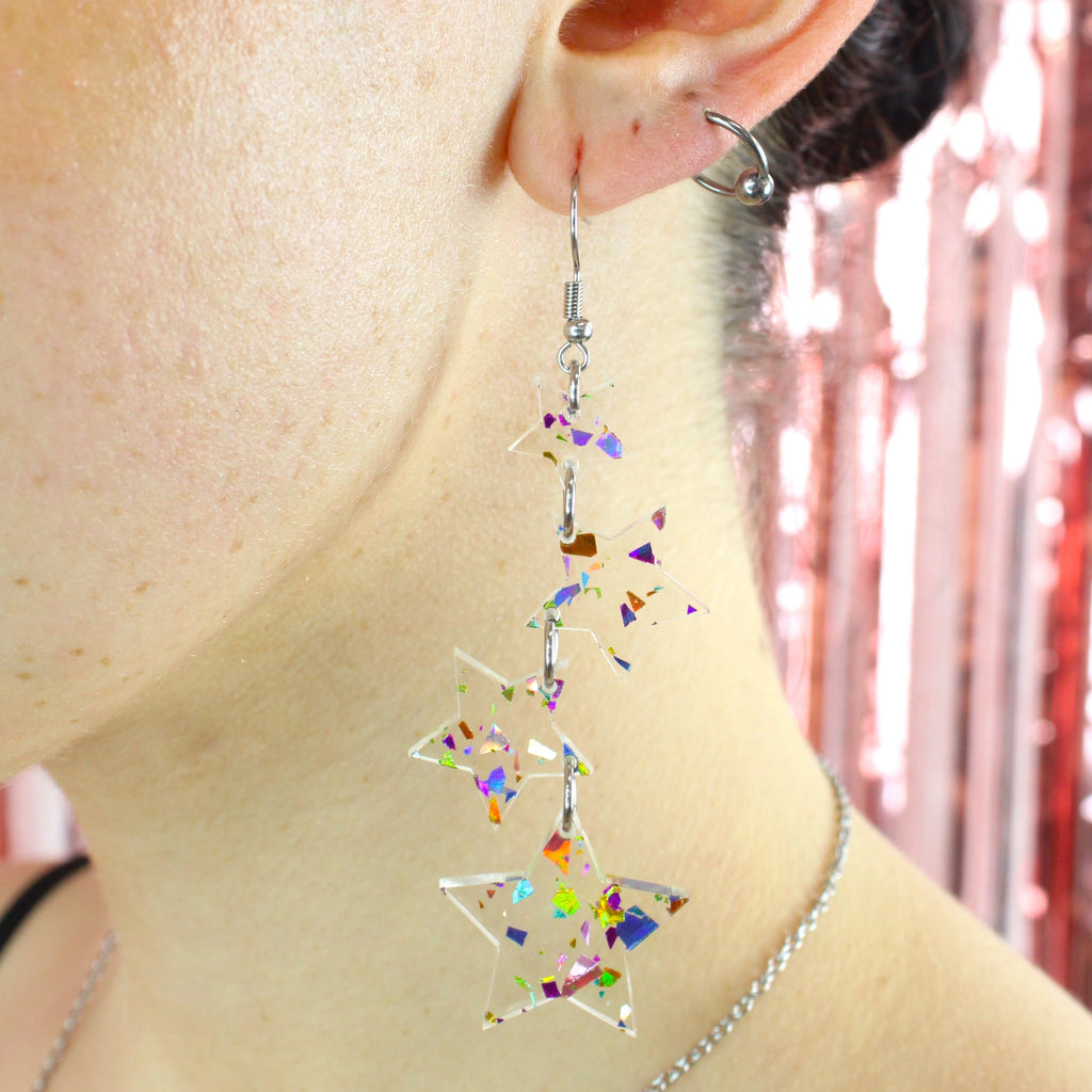 Model wearing four confetti star charms hung from stainless steel earwires ranging in size with a small star at the top followed by two medium stars and a large star at the bottom