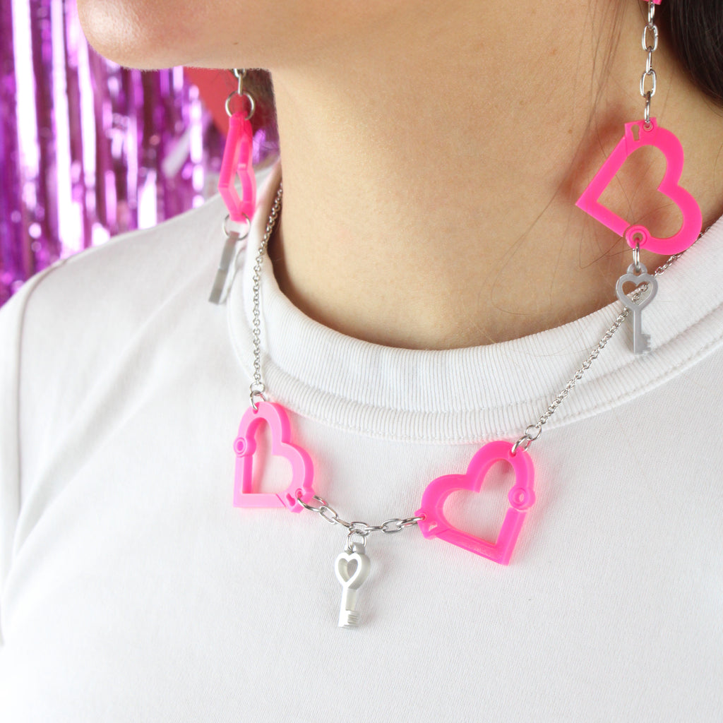 model wearing neon pink acrylic heart charms chained together to resemble handcuffs, with a key charm attached to the bottom. Model is also wearing the matching necklace.