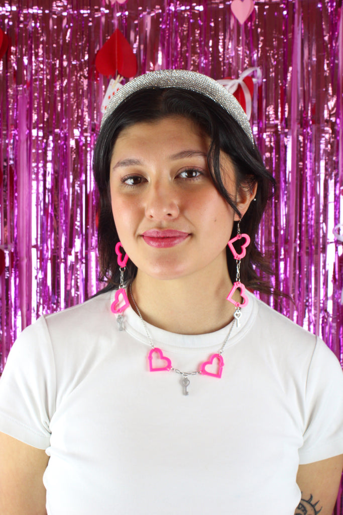 model wearing neon pink acrylic heart charms chained together to resemble handcuffs, with a key charm attached to the bottom. Model is also wearing the matching necklace.