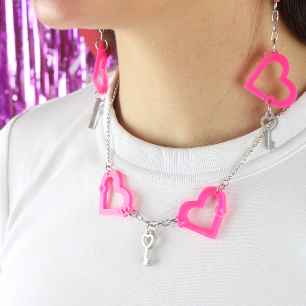 Model wearing neon pink acrylic heart charms chained together to resemble handcuffs, with a key charm in the middle of the two handcuff pendants, on a stainless steel chain. Model is also wearing the matching earrings.