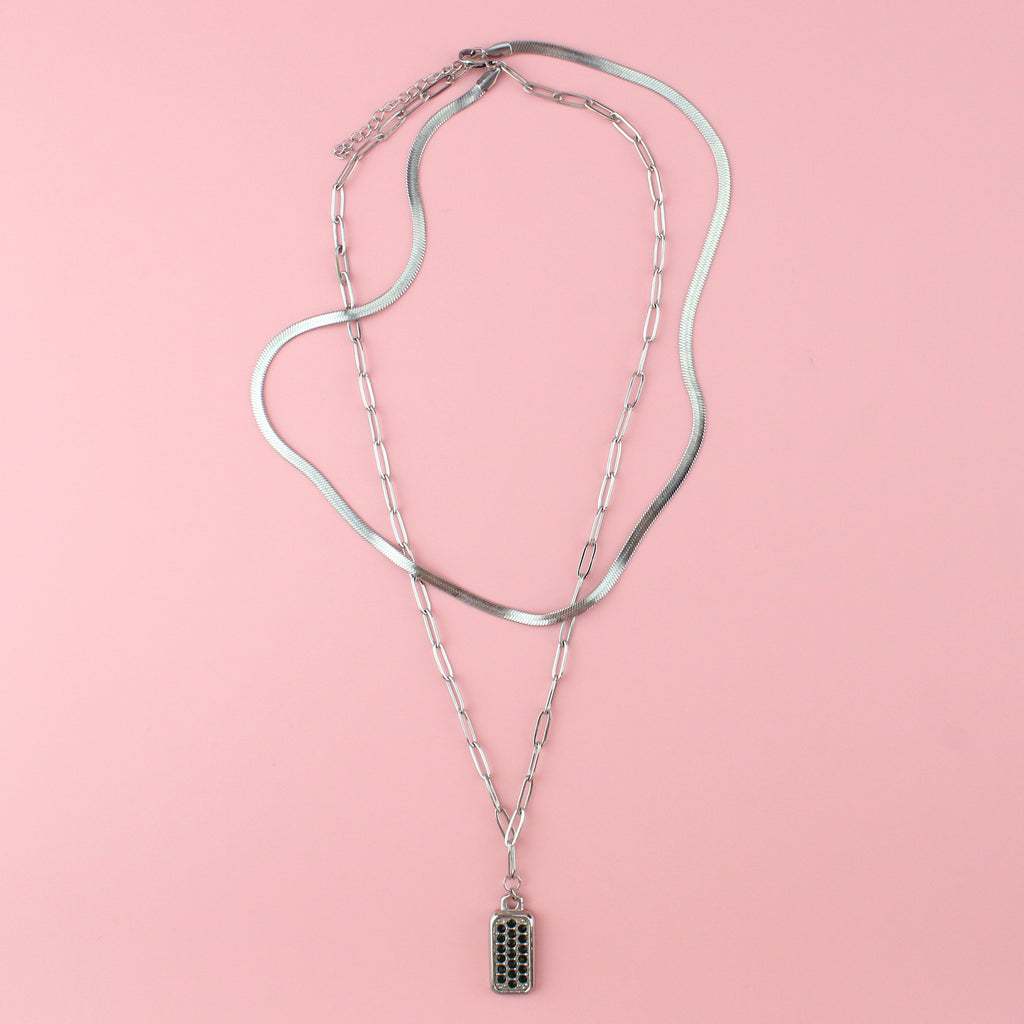A layered necklace featuring both an oval link and snake chain with a rectangular pendant with emerald cubic zirconia stones
