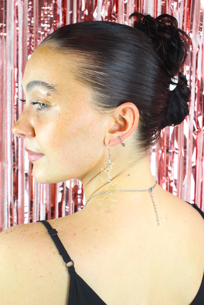 Model wearing Four transparent star charms with gold fleck detailing hanging from stainless steel earwires, varying in size (the largest one hanging at the bottom) 