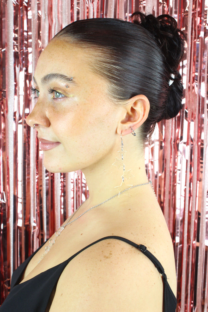 Model wearing Four transparent star charms with gold fleck detailing hanging from stainless steel earwires, varying in size (the biggest one hanging at the bottom)