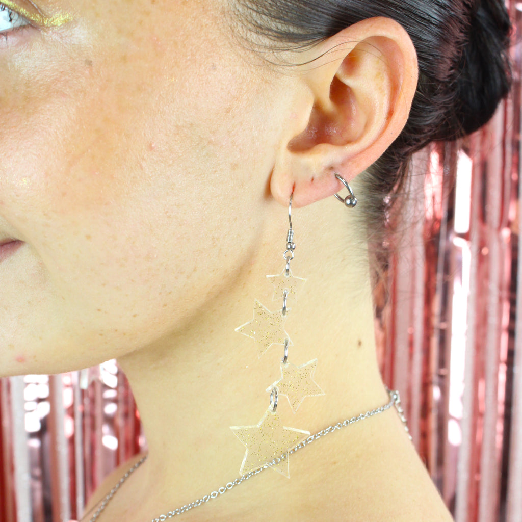 Model wearing four transparent star charms with gold fleck detailing hanging from stainless steel earwires, varying in size (the biggest one hanging at the bottom)