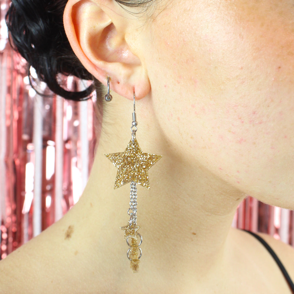 Model wearing a gold glitter acrylic star charm hung from stainless steel earwires with chains dangling from the star that feature a mini star at the end of each of them