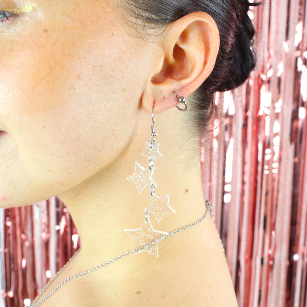 Model wearing four holographic glitter star charms hung from stainless steel earwires ranging in size with a small star at the top followed by two medim stars and a large star at the bottom