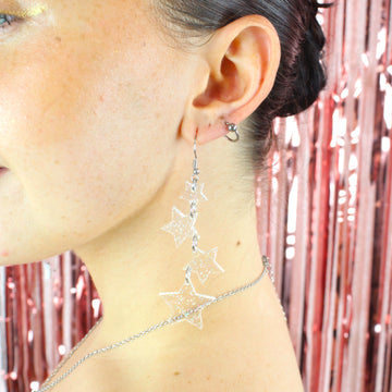 Model wearing four holographic glitter star charms hung from stainless steel earwires ranging in size with a small star at the top followed by two medim stars and a large star at the bottom