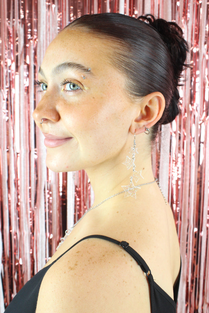 Model wearing four holographic glitter star charms hung from stainless steel earwires ranging in size with a small star at the top followed by two medim stars and a large star at the bottom