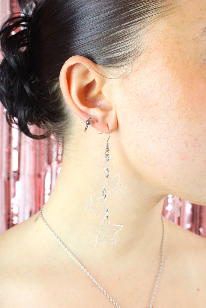 Model wearing four holographic glitter star charms hung from stainless steel earwires ranging in size with a small star at the top followed by two medim stars and a large star at the bottom