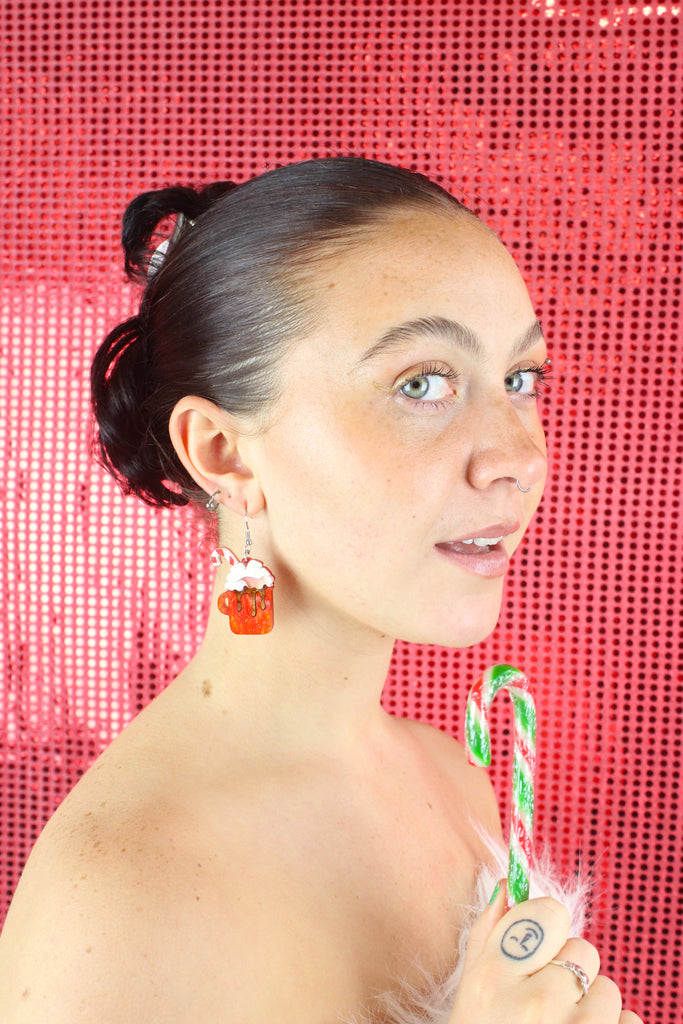 Model wearing Hot chocolate earrings that feature orange glittery mugs with a cloud of whipped cream and a candy cane, hung on stainless steel earwires