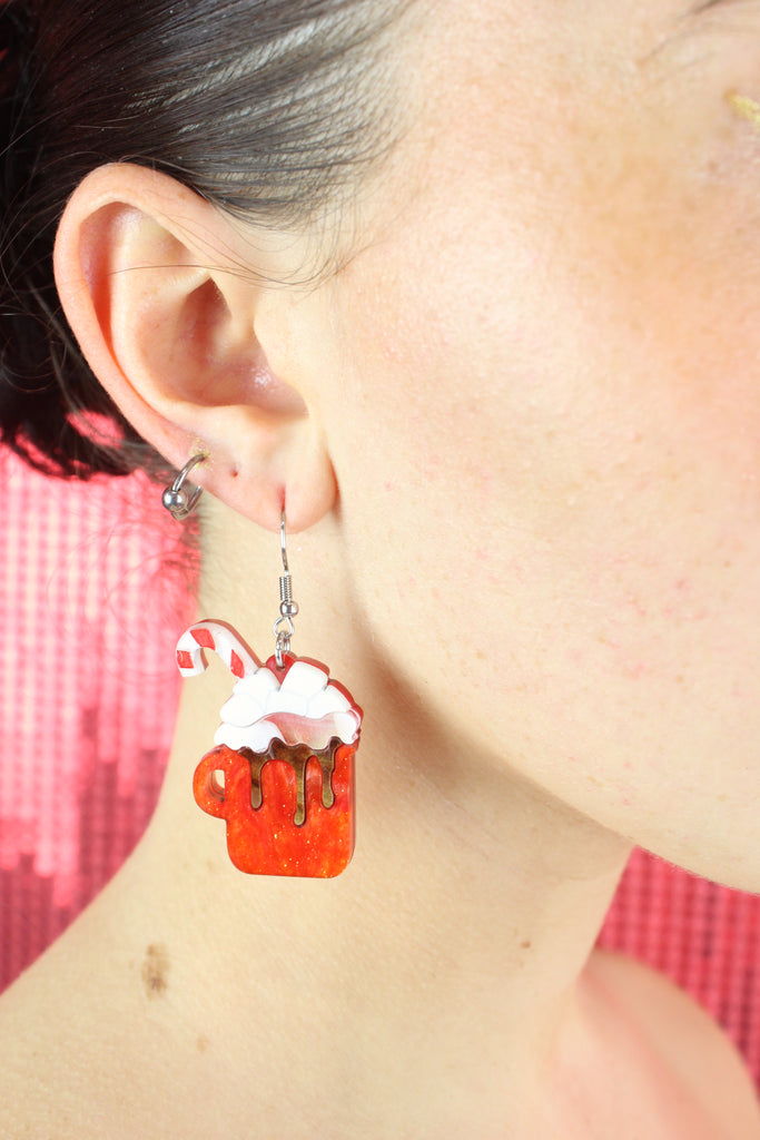 Model wearing Hot chocolate earrings that feature orange glittery mugs with a cloud of whipped cream and a candy cane, hung on stainless steel earwires