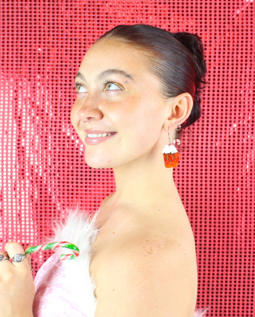 Model wearing Hot chocolate earrings that feature orange glittery mugs with a cloud of whipped cream and a candy cane, hung on stainless steel earwires
