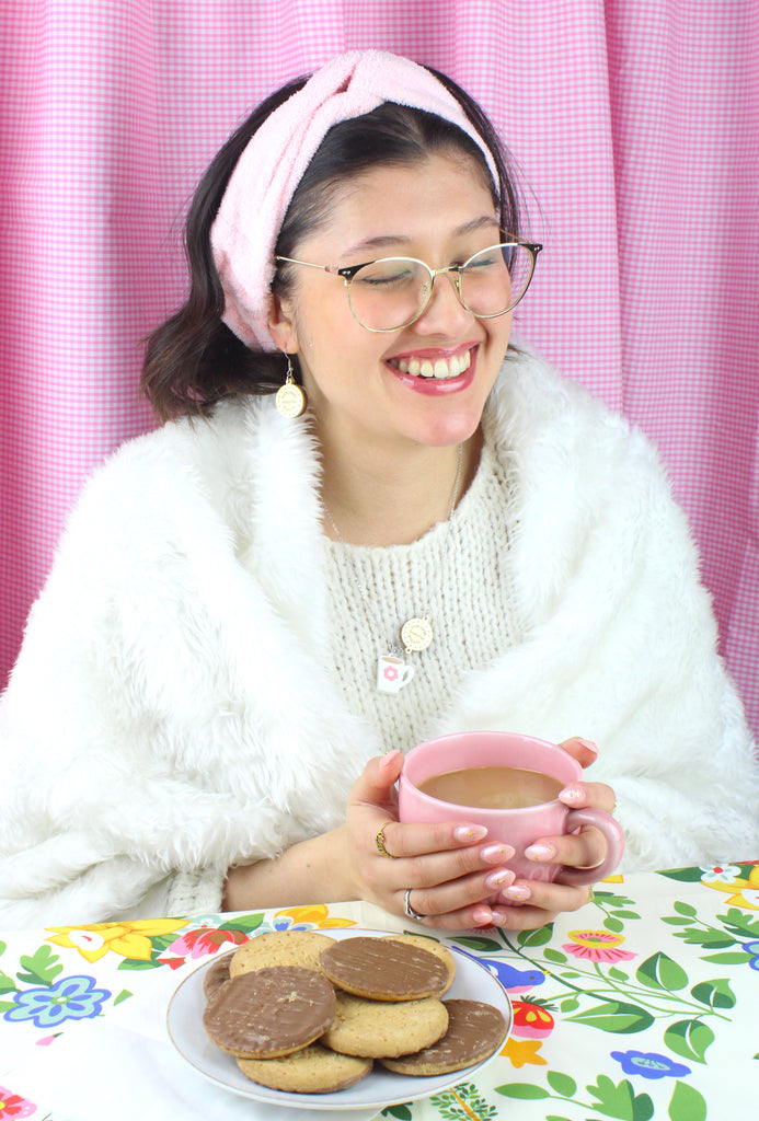 Model wearing Stainless steel chain with a chocolate digestive pendant reading 'Sour Cherry Digestive Biscuits' and a cup of tea pendant featuring a pink flower design, with matching earrings. There is a plate of chocolate digestives in front of her.