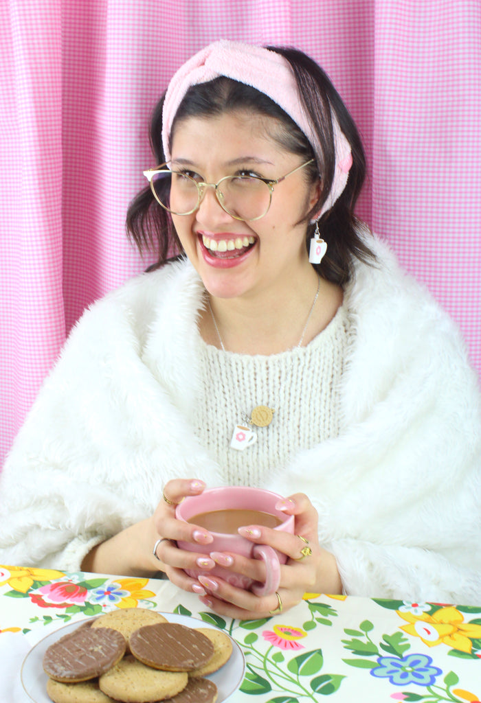 Stainless steel chain with a chocolate digestive pendant reading 'Sour Cherry Digestive Biscuits' and a cup of tea pendant featuring a pink flower design. Model is also wearing matching earrings with a plate of chocolate digestives in front of her.