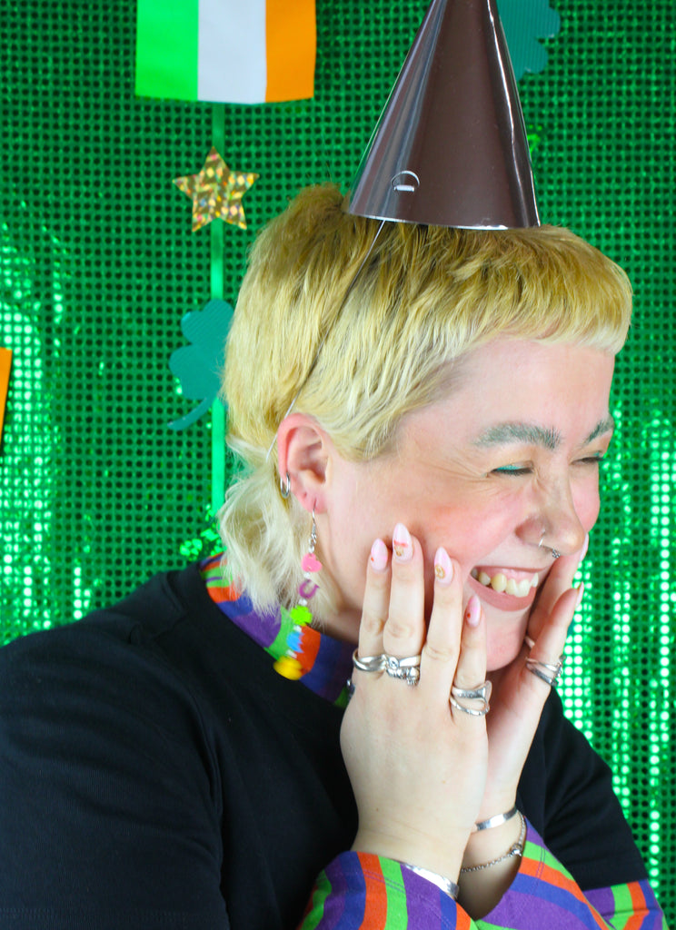 Model wearing a chain of lucky charms featuring a pink heart, a purple horseshoe. a green four leaf clover, a blue moon and orange pot of gold on stainless steel earwires. Model is also wearing a party hat