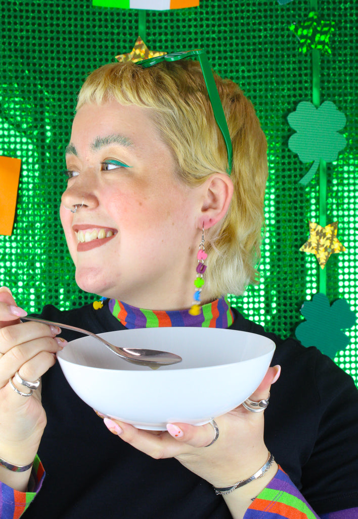 Model wearing a chain of lucky charms featuring a pink heart, a purple horseshoe. a green four leaf clover, a blue moon and orange pot of gold on stainless steel earwires. Model is also holding a bowl of cereal and a spoon