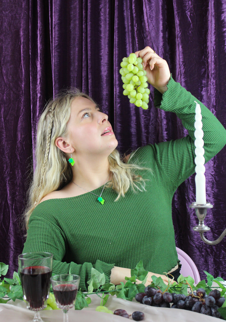 Model wearing stainless steel necklace featuring a bunch of green acrylic grapes on a grapevine, holding a bucnh of green grapes