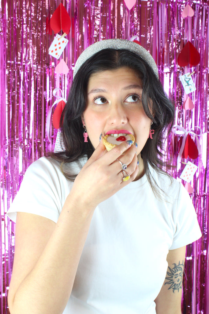 Model wearing mini rose pink bow charms on stainless steel huggie hoops. Model is also eating a cherry bakewell.