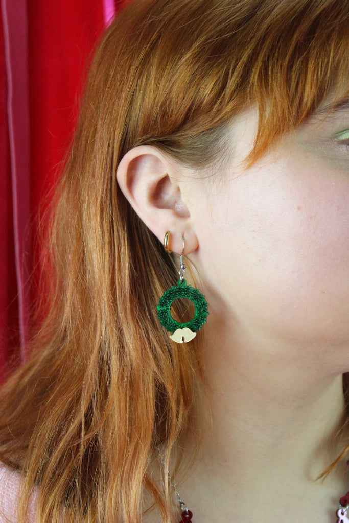 Close-up of a person wearing green wreath earrings with a red curtain background