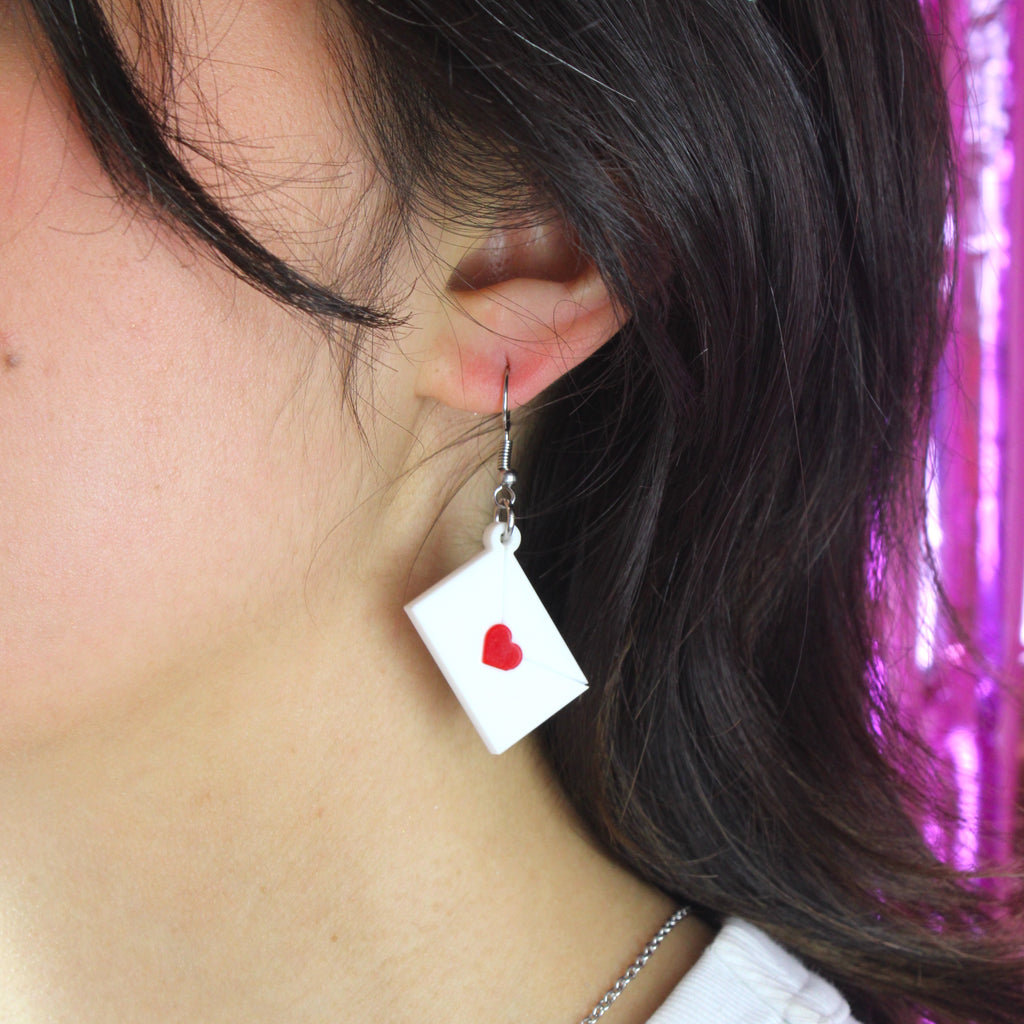 Model wearing white envelope charms sealed with a red heart on stainless steel earwires