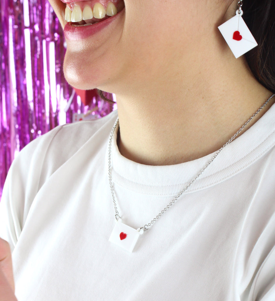 model wearing love letter pendant featuring a white envelope sealed with a red heart on a stainless steel chain. Model is also wearing matching earrings