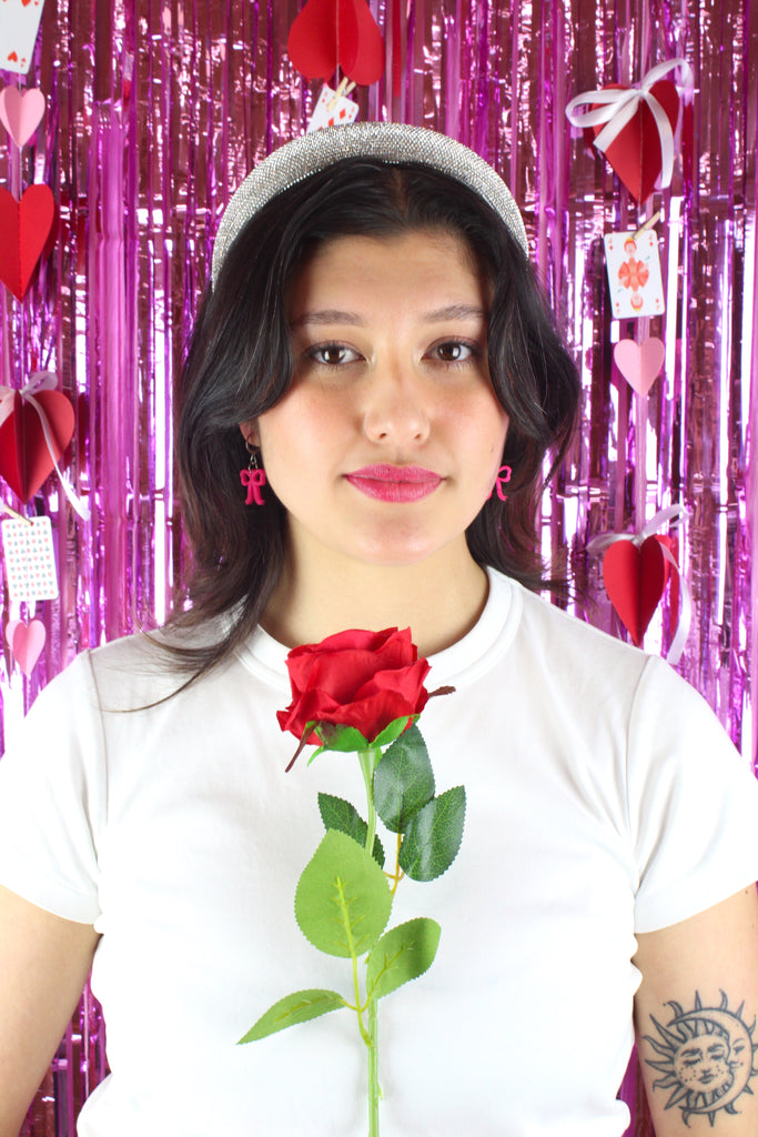 Model wearing Bright pink mini bow charms on stainless steel huggie hoops. Model is also holding a red rose.