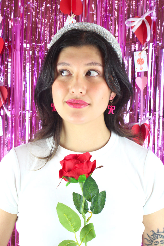 Model wearing Bright pink mini bow charms on stainless steel huggie hoops. Model is also holding a red rose.