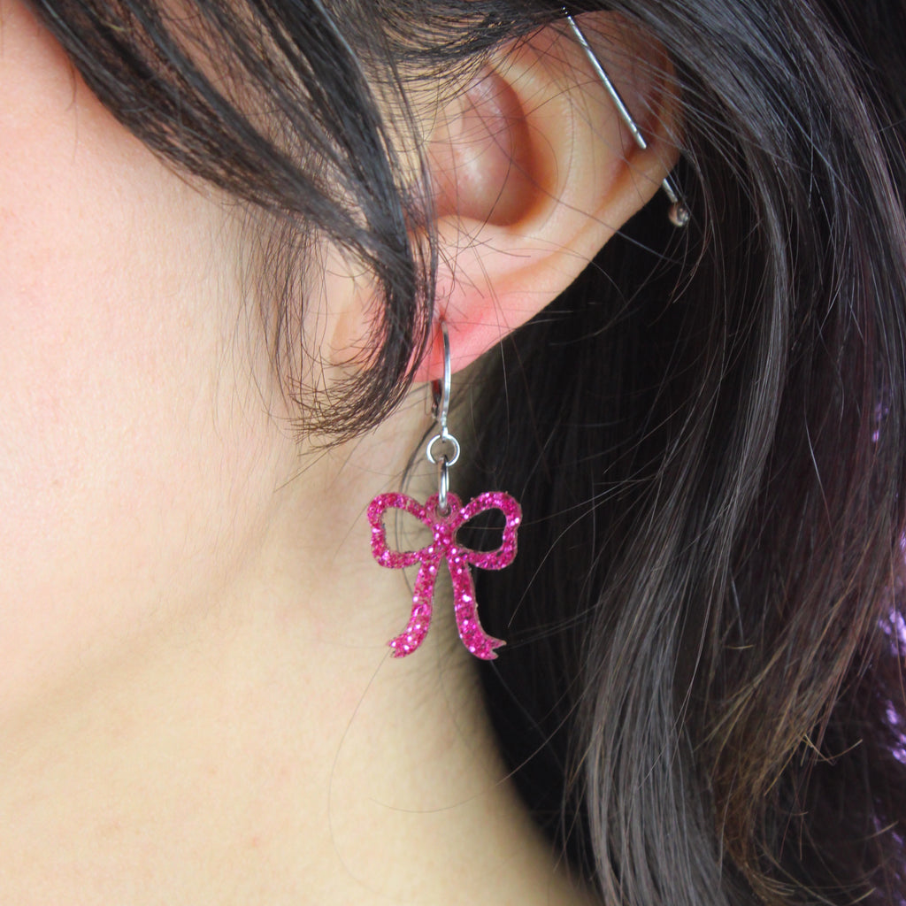Model wearing Mini pink glitter bow charms on stainless steel huggie hoops