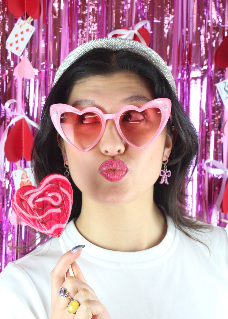 Model wearing Mini pink glitter bow charms on stainless steel huggie hoops. Model is also wearing heart shaped sunglasses and holding a heart-shaped lollipop