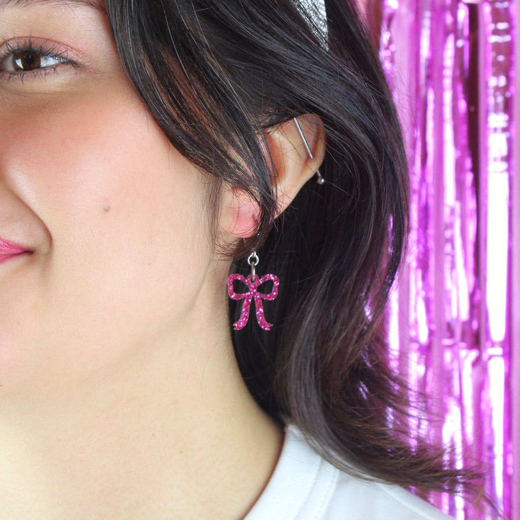 Model wearing Mini pink glitter bow charms on stainless steel huggie hoops. 