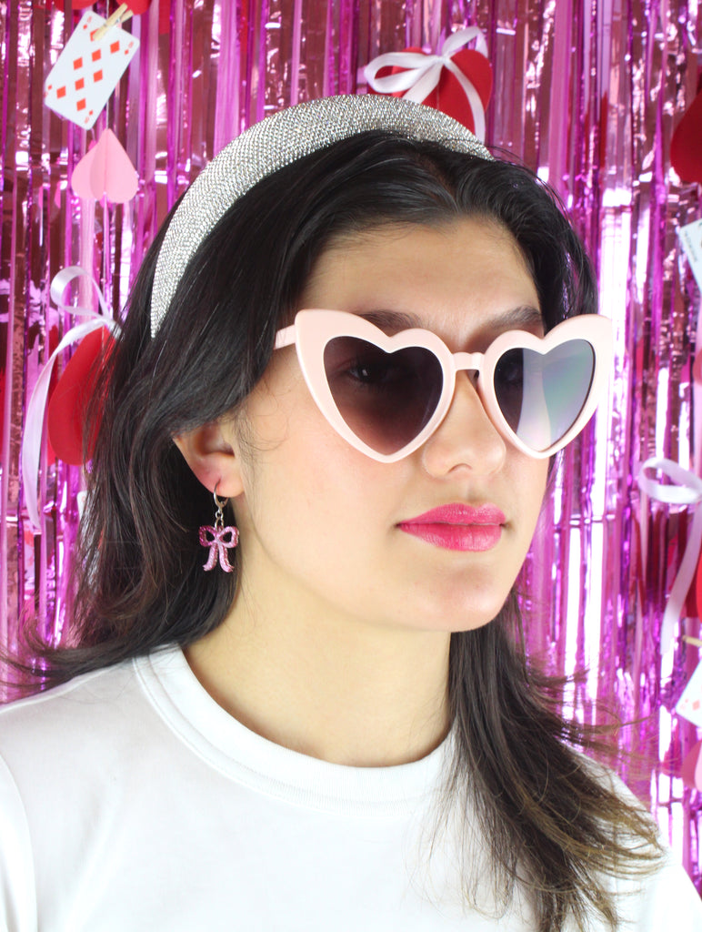 Model wearing Mini pink glitter bow charms on stainless steel huggie hoops. Model is also wearing heart-shaped sunglasses