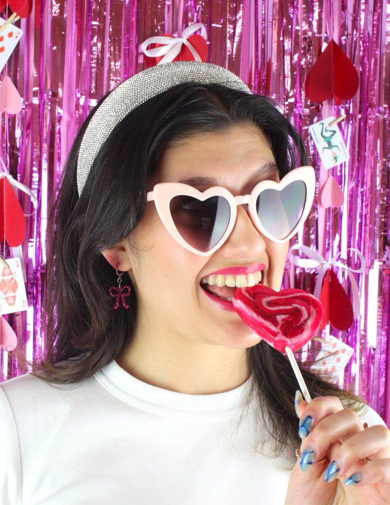 Model wearing Mini pink glitter bow charms on stainless steel huggie hoops. Model is also wearing heart shaped sunglasses and holding a heart-shaped lollipop