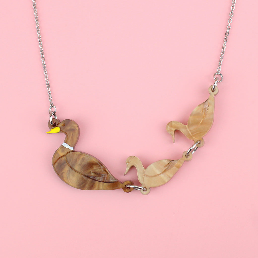 A brown acrylic mother duck pendant with two light brown marble duckling pendants following behind, on a stainless steel chain