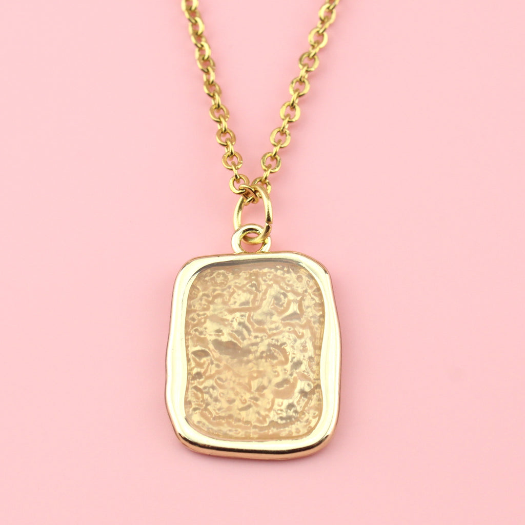 Gold rectangular shaped pendant with a wavy outline and a shimmery peach coloured centre on a gold plated stainless steel chain