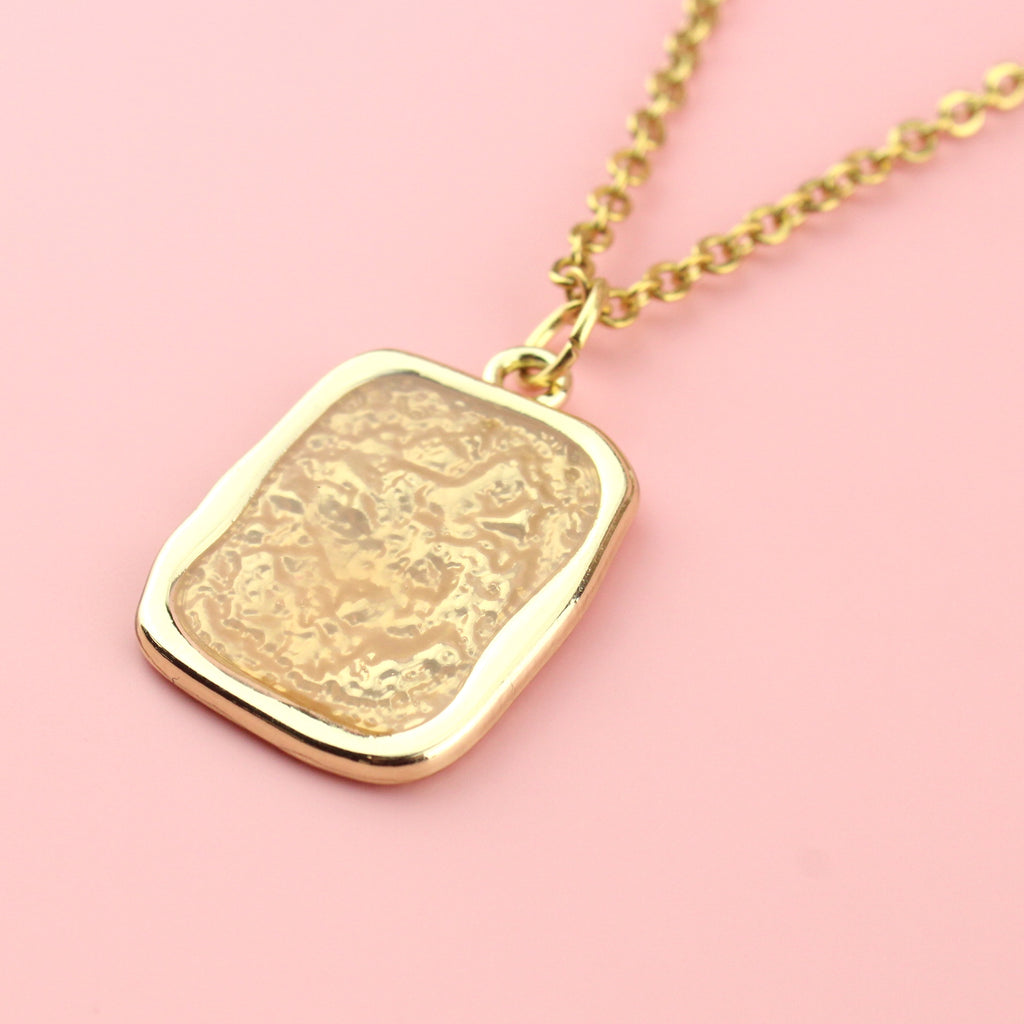 Gold rectangular shaped pendant with a wavy outline and a shimmery peach coloured centre on a gold plated stainless steel chain