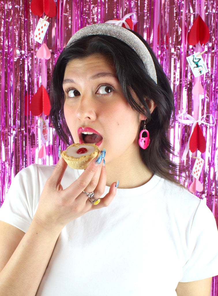 Model wearing pink heart padlock earring , holding a cherry bakewell tart