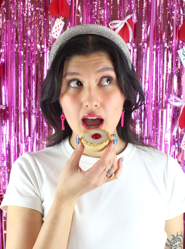 Model wearing pink heart padlock earring and key earring holding a cherry bakewell tart