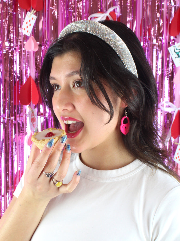 Model wearing pink heart padlock earring , holding a cherry bakewell tart