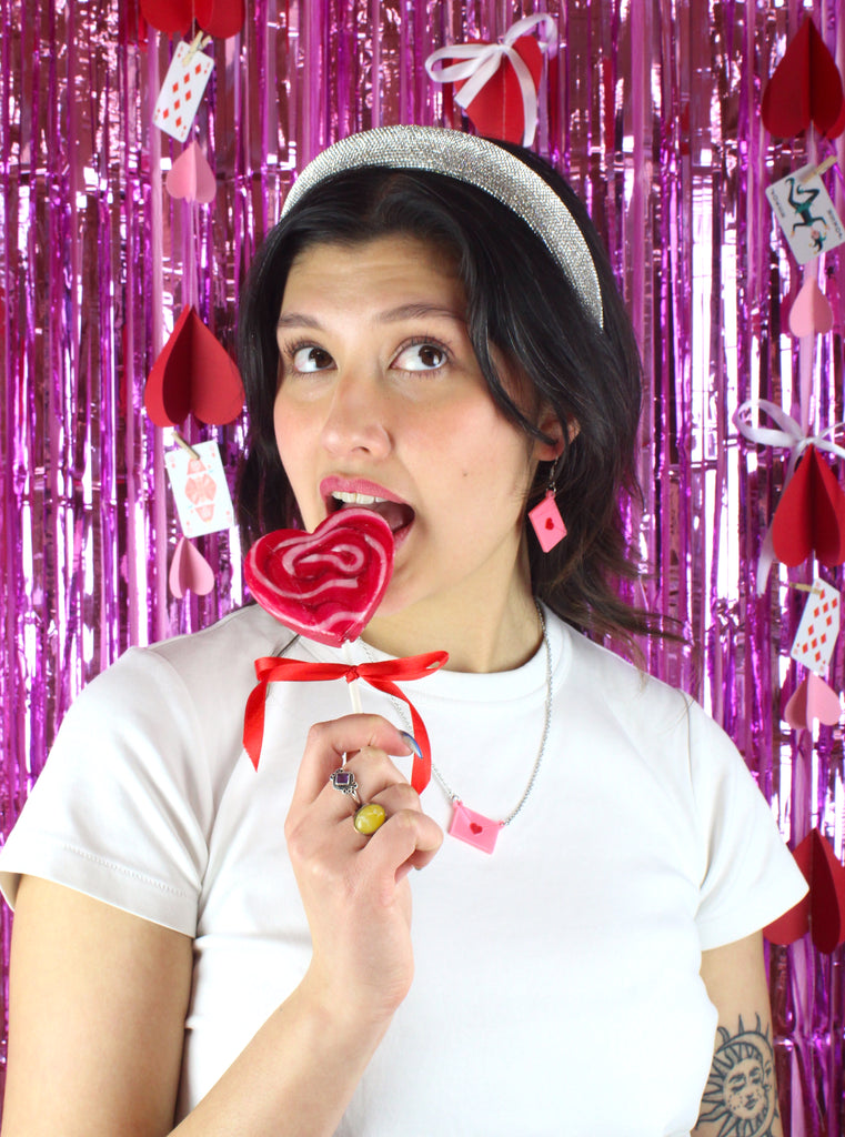 Model wearing acrylic pink envelopes, each sealed with a red heart, and attached to stainless steel earwires. Model is also wearing matching necklace and holding a heart shaped lollipop