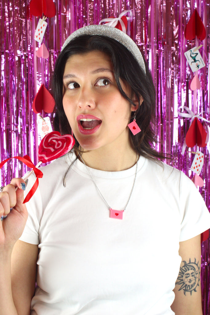Model wearing acrylic pink envelopes, each sealed with a red heart, and attached to stainless steel earwires. Model is also wearing matching necklace and holding a heart shaped lollipop