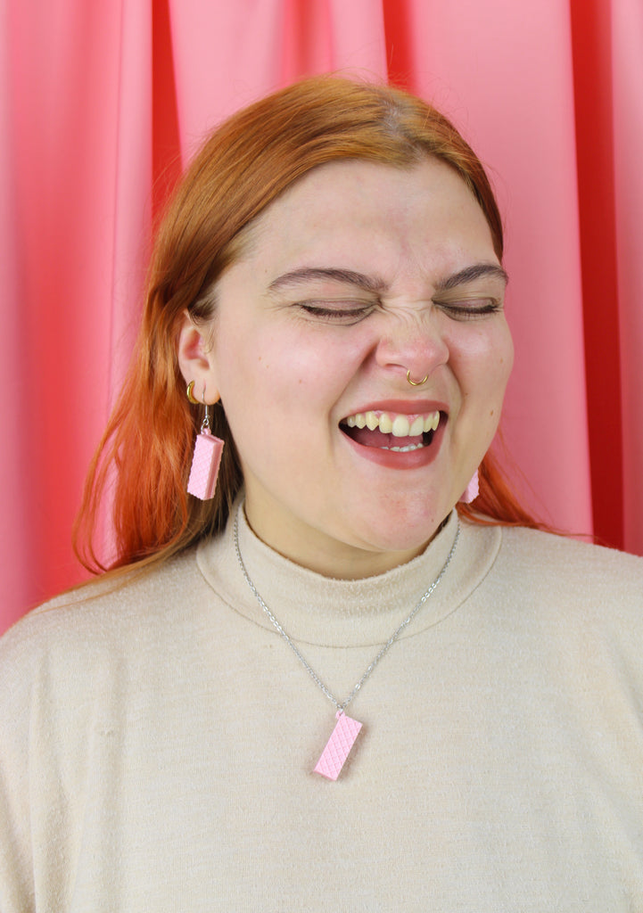 Person with red hair wearing pink earrings and a necklace against a pink curtain background