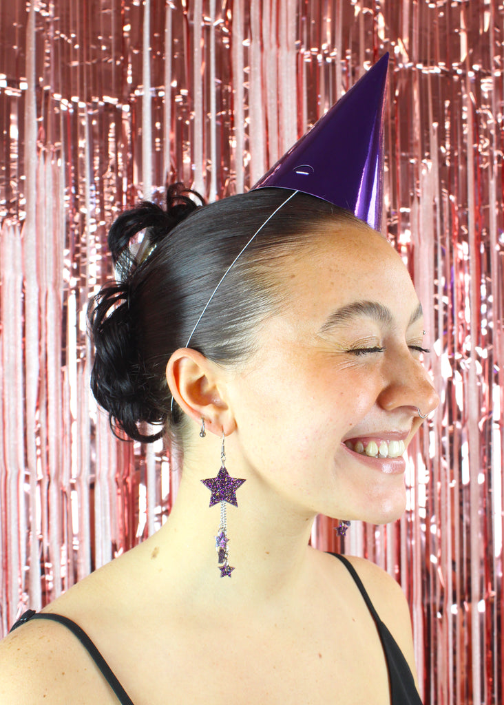 Purple glitter acrylic stars hanging from stainless steel earwires with three chains hanging from the star with mini stars at the end of each chain. Model is wearing a party hat