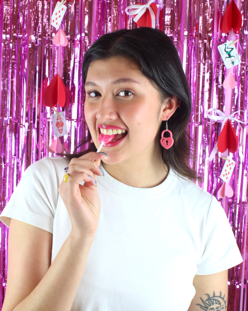 Model wearing Rose pink heart padlock charm on a stainless steel earwire, biting a pink lollipop